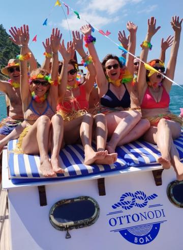 Gruppo festoso in barca, sorrisi e braccia alzate, atmosfera di gioia e libertà.